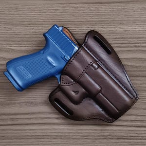 Puede incluir: Una funda de cuero marrón oscuro con costuras visibles y dos ranuras para cinturón, que contiene una pistola de polímero azul brillante. La funda presenta una forma moldeada para ajustarse al arma, con un acabado liso y forro oscuro visible.