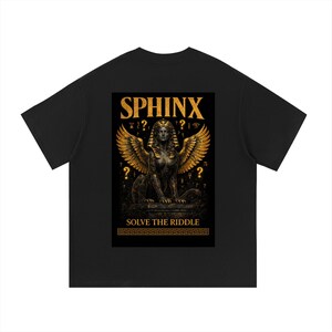 以下が含まれることがあります： 黒いTシャツ。金色の翼を持つスフィンクスのグラフィックと、その上に「SPHINX」の文字。画像の下には「SOLVE THE RIDDLE」のフレーズ。デザインには疑問符と古代エジプトのシンボルが含まれています。