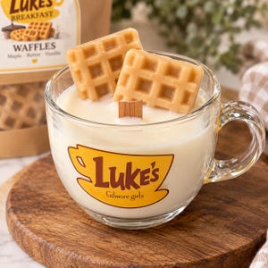 May include: A clear glass mug filled with white wax and a wooden wick, topped with two tan waffle-shaped wax pieces. The mug features a yellow graphic with the brown text 'Luke's' and 'Gilmore girls'. A matching bag is visible in the background.