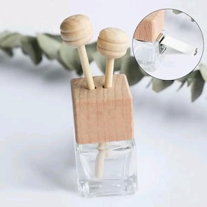 May include: A clear glass square bottle with a light wood cap featuring two wooden honey dipper sticks inserted into the top. A circular inset shows a metal clip attached to the side of the wooden cap, designed for mounting the bottle.