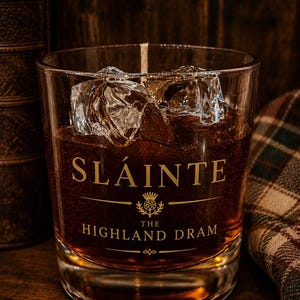 May include: A clear glass candle vessel filled with amber-coloured wax, featuring the gold-printed text SLÁINTE THE HIGHLAND DRAM and a thistle emblem. The candle contains realistic-looking clear wax ice cubes and a single centred cotton wick.