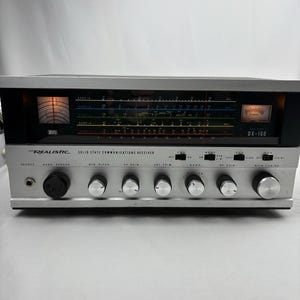 Puede incluir: Un receptor de comunicaciones de estado sólido Realistic DX-160 plateado. La parte frontal tiene una serie de perillas, diales e interruptores. La parte superior tiene una pantalla con un dial de sintonización y una escala de frecuencia. El texto "Realistic" y "DX-160" son visibles.