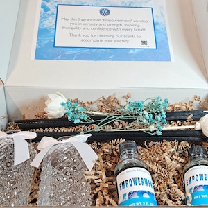 May include: A white gift box with a blue and white card that says "May this fragrance of 'Empowerment' envelop you in serenity and strength, inspiring tranquility and confidence with every breath. Thank you for choosing our scents to accompany your journey." The box contains two clear glass bottles with black labels that say "EMPOWERMENT" and have a blue and white design. The bottles are surrounded by brown packing paper and white and blue dried flowers.