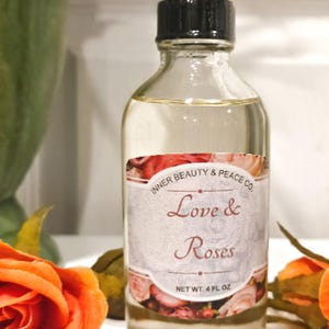 May include: A clear glass bottle with a black cap containing a light-colored liquid. The label features a floral pattern with the text Inner Beauty & Peace Co. Love & Roses and Net Wt. 4 fl oz. The bottle is positioned between two orange roses.