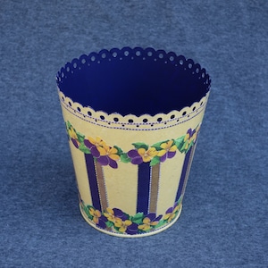 May include: A yellow metal wastebasket with a blue interior and a scalloped rim. The wastebasket is decorated with a floral pattern of purple and yellow flowers.
