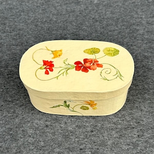 May include: A white oval box with a floral design featuring red and yellow flowers and green leaves. The box is made of wood and has a smooth finish.