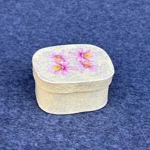May include: A small, cream-colored box with a floral design. The box has a textured surface and features pink flowers with white centers.