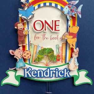 May include: A layered paper cake topper featuring a rainbow arch, the text ONE for the books, and the name Kendrick. It includes illustrations of The Cat in the Hat, Curious George, Winnie the Pooh, Peter Rabbit, and The Very Hungry Caterpillar.
