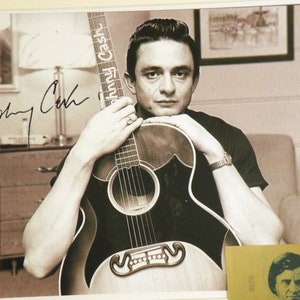 K&ouml;nnte beinhalten: Schwarzwei&szlig;foto von Johnny Cash, der eine Gitarre h&auml;lt. Die Gitarre hat einen schwarzen Korpus und ein wei&szlig;es Schlagbrett. Die Gitarre hat einen Aufkleber am Hals, auf dem "Johnny Cash" steht. Das Foto ist signiert mit "Johnny Cash".