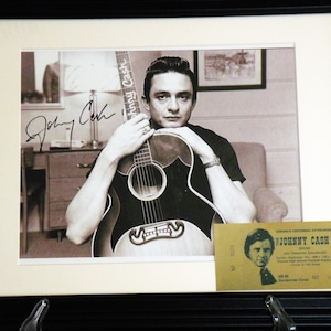 K&ouml;nnte beinhalten: Ein Schwarzwei&szlig;foto von Johnny Cash, der eine Gitarre h&auml;lt. Das Foto ist mit "Johnny Cash" in schwarzer Tinte signiert. Ein gelbes Ticket f&uuml;r ein Johnny Cash Konzert befindet sich in der rechten unteren Ecke des Bildes.