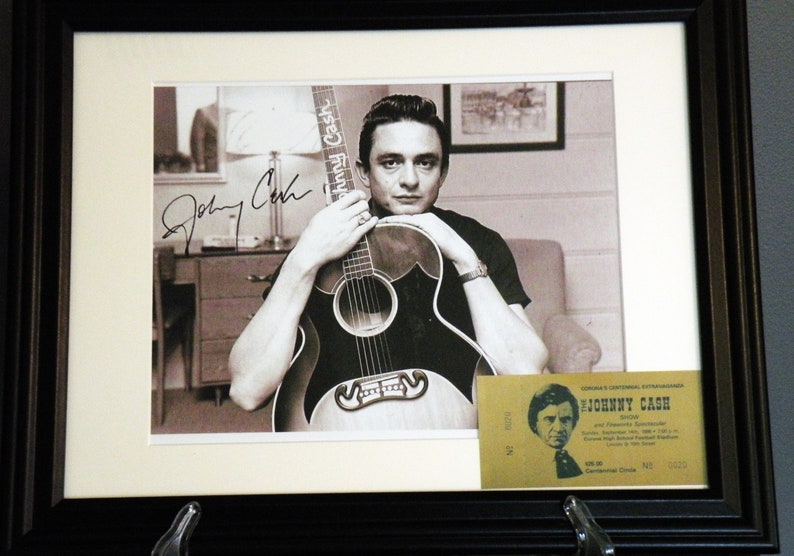 K&ouml;nnte beinhalten: Ein Schwarzwei&szlig;foto von Johnny Cash, der eine Gitarre h&auml;lt. Das Foto ist mit "Johnny Cash" in schwarzer Tinte signiert. Ein gelbes Ticket f&uuml;r ein Johnny Cash Konzert befindet sich in der rechten unteren Ecke des Bildes.