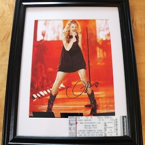 May include: A framed photograph of a woman singing on stage. The woman is wearing a black dress and cowboy boots. The photo is signed with a black marker. The photo is accompanied by a ticket stub for a Taylor Swift concert at Lincoln Financial Field on July 20, 2013.