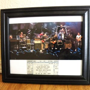 Op de afbeelding: Een zwarte rechthoekige lijst met een foto van de Allman Brothers Band tijdens een optreden in het Red Rocks Amphitheatre, geplaatst boven een concertticket voor het evenement op zaterdag 30 augustus 2008 om 19:00 uur.