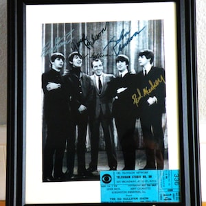 Puede incluir: Una fotografía en blanco y negro de The Beatles firmada por los cuatro miembros. La foto muestra a los miembros de la banda de pie frente a una cortina con un boleto de CBS Television Network para The Ed Sullivan Show en la esquina inferior derecha.