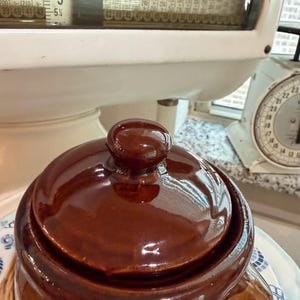 May include: A dark brown glazed ceramic crock with a matching lid featuring a central knob handle. The crock has a smooth, reflective surface and sits on a patterned plate. The background shows a white vintage scale with visible numerical charts.