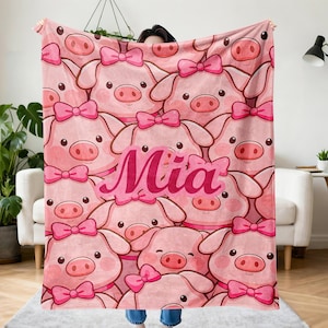 May include: Pink blanket featuring a pattern of cartoon pigs with pink bows. The name "Mia" is displayed in pink script. The blanket is a soft, cozy item, perfect for a child's room or as a gift.