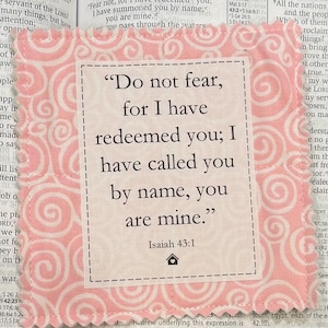 May include: A square, fabric coaster with a pink and white swirl pattern border. The center features a cream-colored square with the text "Do not fear, for I have redeemed you; I have called you by name, you are mine." Isaiah 43:1.