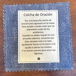 May include: A small, square prayer quilt with a blue patterned fabric border and a white center. The center features Spanish text. The quilt is designed to offer comfort and support during difficult times. The text encourages faith and resilience.