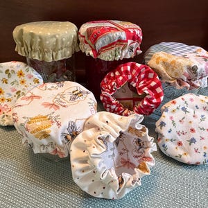 Op de afbeelding: Een collectie stoffen potdeksels met diverse patronen, waaronder bijen, bloemenprints, rood-witte ruitjes en stippen. Deze ronde, elastische textieldeksels zijn ontworpen om over glazen potten te passen en tonen een verscheidenheid aan kleuren.