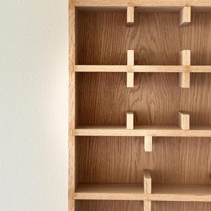 Puede incluir: Una estanter&iacute;a de madera con m&uacute;ltiples estantes horizontales y soportes verticales. Los estantes son de madera clara y est&aacute;n unidos a los soportes verticales con peque&ntilde;os bloques de madera. El fondo es una pared blanca.