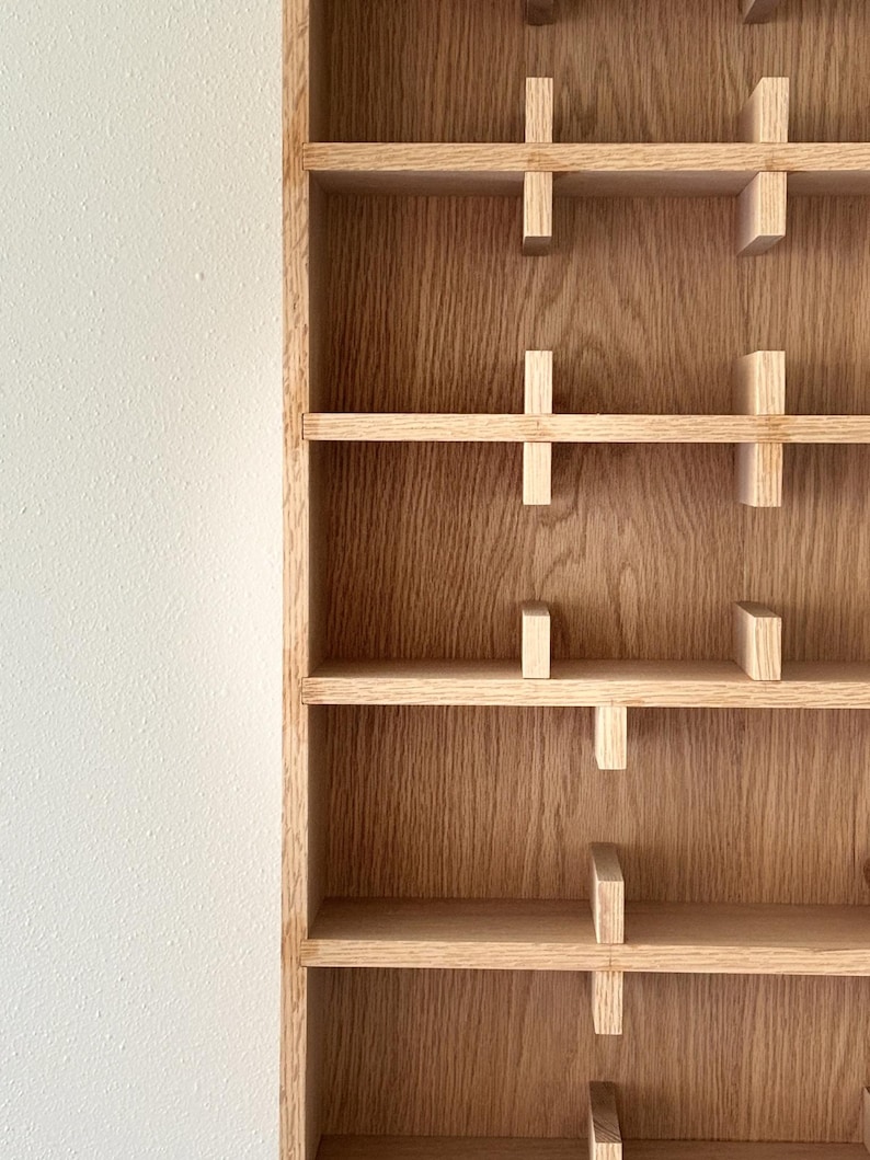 Puede incluir: Una estanter&iacute;a de madera con m&uacute;ltiples estantes horizontales y soportes verticales. Los estantes son de madera clara y est&aacute;n unidos a los soportes verticales con peque&ntilde;os bloques de madera. El fondo es una pared blanca.