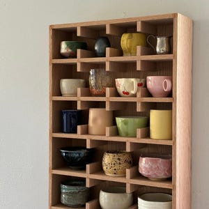 Puede incluir: Una estanter&iacute;a de madera con m&uacute;ltiples compartimentos, cada uno con una taza o cuenco de cer&aacute;mica &uacute;nico. Las tazas var&iacute;an en color, incluyendo amarillo, rosa, azul y verde, y presentan diversos dise&ntilde;os y texturas. La estanter&iacute;a est&aacute; montada en una pared gris claro.