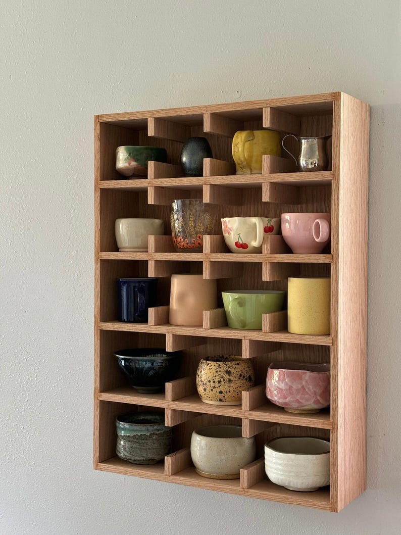 Puede incluir: Una estanter&iacute;a de madera con m&uacute;ltiples compartimentos, cada uno con una taza o cuenco de cer&aacute;mica &uacute;nico. Las tazas var&iacute;an en color, incluyendo amarillo, rosa, azul y verde, y presentan diversos dise&ntilde;os y texturas. La estanter&iacute;a est&aacute; montada en una pared gris claro.