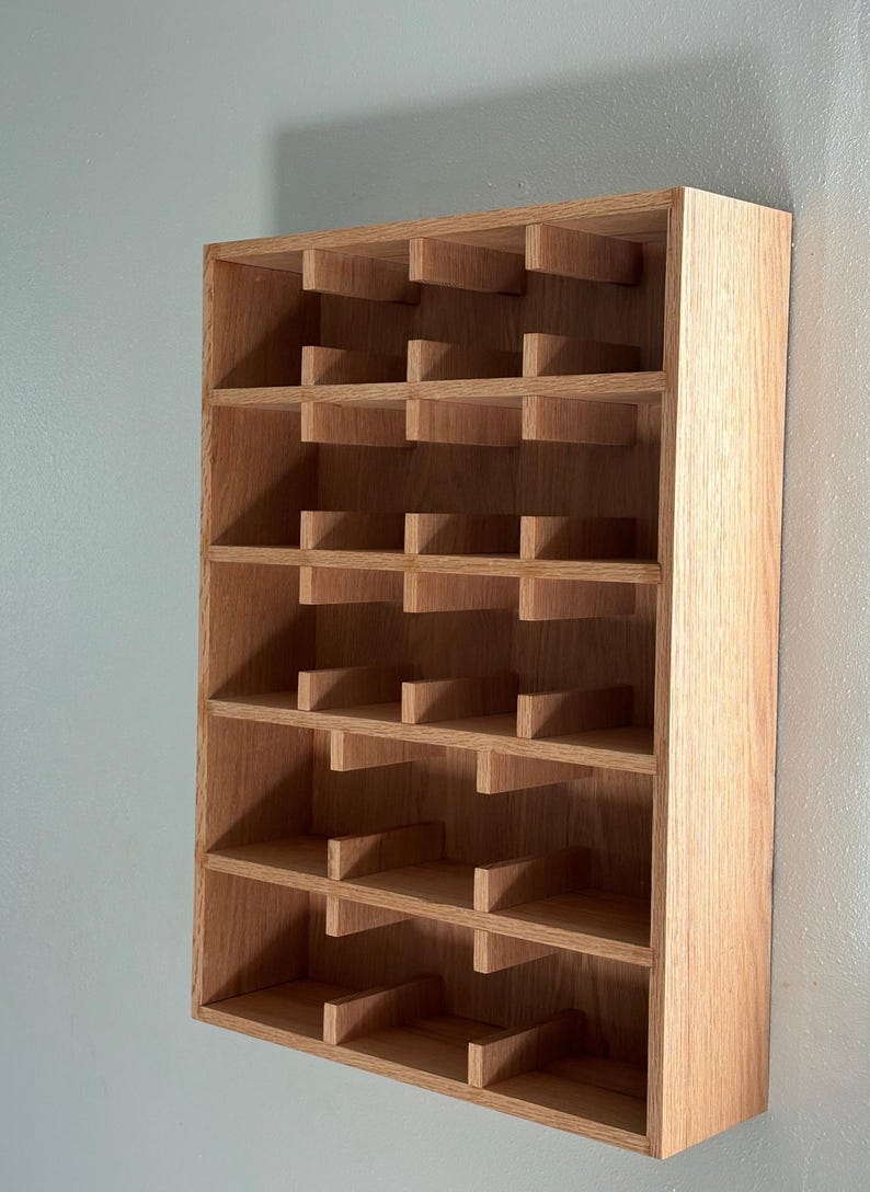 Puede incluir: Un botellero de madera para montaje en pared con m&uacute;ltiples compartimentos. El botellero es de madera clara y tiene forma rectangular. Est&aacute; dise&ntilde;ado para guardar botellas de vino, con cada compartimento dimensionado para una sola botella. Est&aacute; montado en una pared azul claro.