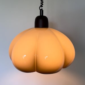 Op de afbeelding: Een hanglamp met een glanzende, lichtgele, pompoenvormige glazen kap met een glad, afgerond oppervlak. De kap is bevestigd aan een zwarte cilindrische dop en hangt aan een zwart spiraalsnoer dat naar boven toe loopt.