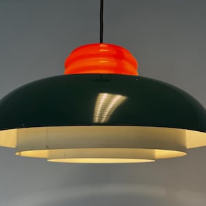 Op de afbeelding: Een moderne hanglamp met een donkergroene, koepelvormige kap en een feloranje bovenkant. De lamp heeft een gelaagd ontwerp met een wit interieur en hangt aan een zwart snoer. De armatuur is verlicht.
