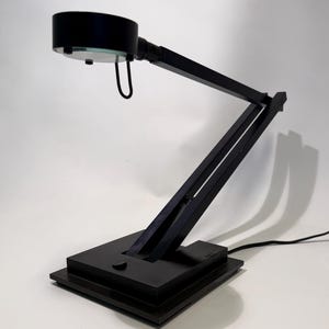 May include: A black desk lamp with an adjustable arm and a circular lamp head. The lamp has a rectangular base and a sleek, modern design. The lamp is plugged into a wall outlet.