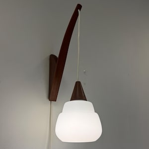 Può includere: Applique da parete in stile moderno con un braccio in legno scuro e un paralume in vetro bianco. La lampada è montata su una parete grigia e ha un cavo bianco. Il design è minimalista ed elegante.