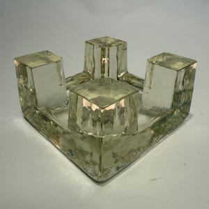 May include: A clear glass candle holder with a textured surface. The holder has a square base with four cube-shaped candle holders. The glass has a slight yellow tint.