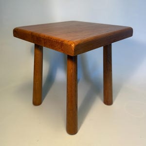 May include: A small, square wooden stool with a warm, medium-brown finish. The stool features a flat, solid top with visible wood grain patterns and four sturdy, cylindrical legs that taper slightly towards the bottom, providing a stable base.