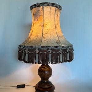 Op de afbeelding: Een vintage tafellamp met een houten voet en een textuur lampenkap. De beige lampenkap heeft bruine accenten en een decoratieve franje. De lamp heeft een klassiek ontwerp, ideaal om een warme gloed aan elke kamer toe te voegen. Ongeveer 60 cm hoog.