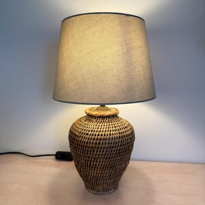 Op de afbeelding: Een geweven rotan tafellamp met een beige stoffen lampenkap. De lampvoet heeft een bolvorm met een geweven textuur. De lampenkap is neutraal van kleur en de lamp is ingeschakeld en geeft een warme gloed.