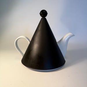 Könnte beinhalten: Ein schwarzer, kegelförmiger Teekannen-Deckel mit einem kleinen, runden Knopf oben. Der Deckel sitzt auf einer weißen Teekanne mit gebogenem Griff und Ausguss. Das Design ist minimalistisch und modern.