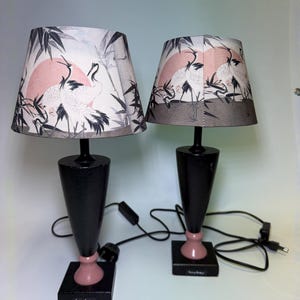 Op de afbeelding: Twee bijpassende tafellampen met zwarte voetstukken en roze accenten. De lampenkappen tonen een Japans geïnspireerd ontwerp met kranen, bamboe en een roze zon tegen een grijze en witte achtergrond. Elke lamp heeft een zwart snoer en stekker.