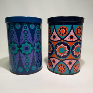 Op de afbeelding: Twee cilindrische metalen blikken met deksels. Eén is marineblauw met een paars en blauwgroen geometrisch patroon. De andere is turquoise, roze en oranje met een stervormig ontwerp. Beide hebben marineblauwe deksels.