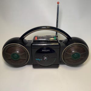 Op de afbeelding: Zwarte Philips Roller 2 boombox met een zilveren antenne en handvat. De radio heeft twee luidsprekers met gaasafdekkingen en een centraal bedieningspaneel. De radio heeft de tekst "Philips" en "Roller 2".