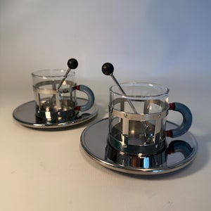 Op de afbeelding: Twee doorzichtige glazen theekopjes met metalen frames en blauwe handvatten staan op bijpassende zilveren schotels. Elke kop heeft een kleine zilveren lepel met een zwarte bal erop. De kopjes zijn ontworpen voor thee of koffie.