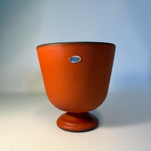 Op de afbeelding: Een gebrande oranje keramische kom op voet met een matte, gespikkelde textuur. De kom heeft een brede, uitlopende opening die taps toeloopt naar een ronde voet. Een kleine, ovale zilverkleurige sticker is bevestigd op de bovenkant aan de voorzijde.