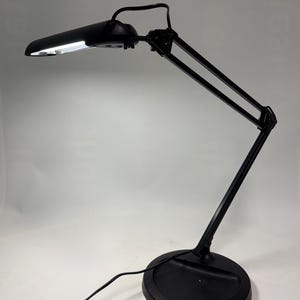 May include: A black desk lamp with an adjustable arm and a round base. The lamp has a rectangular light fixture and a power cord. The light is on, illuminating the area below. The lamp is designed for task lighting.