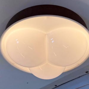 Op de afbeelding: Een moderne plafondlamp met een donkerbruine basis en een wit ontwerp met meerdere lampen. De lamp heeft een unieke vorm, met drie ronde, gloeiende lampen. De armatuur is op een wit plafond gemonteerd.