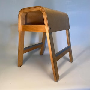 以下が含まれることがあります： 積層合板で作られた、湾曲したサドル型の座面を持つ明るい色の木製スツール。4本の頑丈な脚が水平な支持バーで連結された構造で、木材の自然な木目と質感がはっきりと見て取れます。