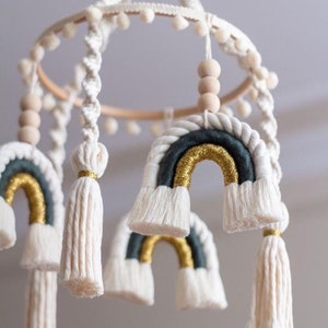 Könnte beinhalten: Ein weißes und goldenes Makramee-Regenbogen-Mobile mit Holzperlen und weißen Pompons. Das Mobile ist aus Baumwollseil gefertigt und hat einen Holzring.
