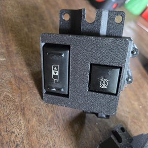 May include: A black rectangular automotive switch panel with a textured finish, featuring two integrated buttons. The left button displays a window icon with up and down arrows, while the right button shows a circular icon with a central symbol.