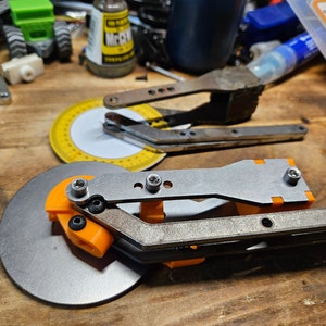 May include: A collection of tools and hardware components on a wooden surface. The items include a circular metal cutter with orange accents, a protractor, and various metal tools. The scene suggests a workshop or a DIY project.