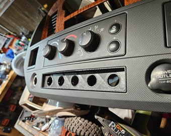 May include: A gray car dashboard component with black control knobs, buttons, and a power outlet. The dashboard has a textured surface and a wood-grain trim.