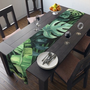 May include: A green and black tropical leaf patterned table runner on a dark wood table with two chairs and two wine glasses.
